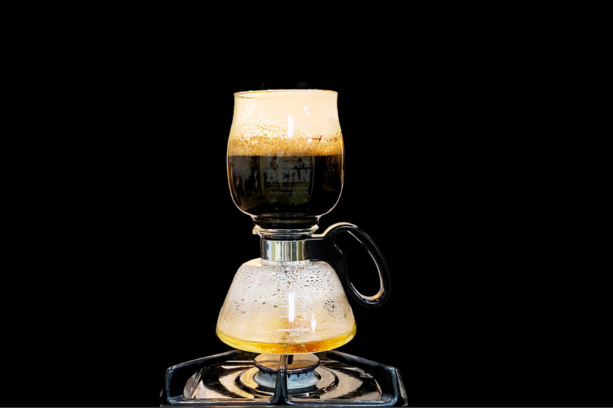 A Yama SY-5 siphon brewing against a black background, coffee steeping in the upper globe above the burner, the I Have a Bean logo showing through the glass