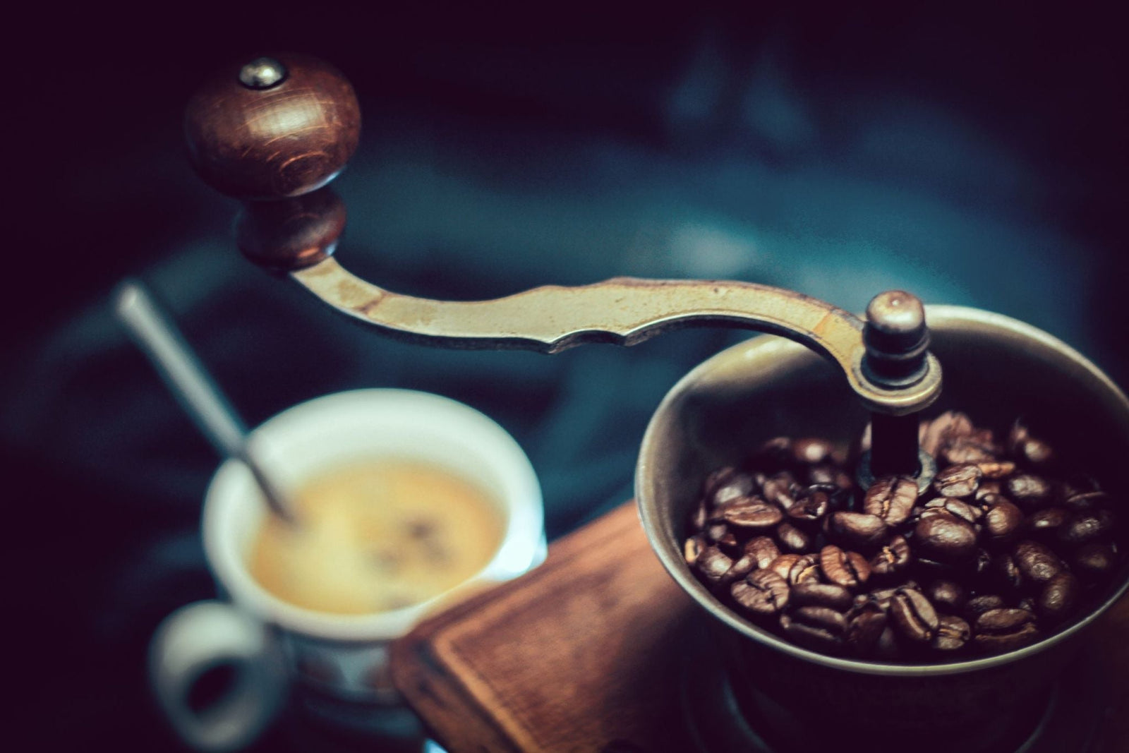 old fashioned hand grinder full of coffee beans. An out of focus espresso is in the background.