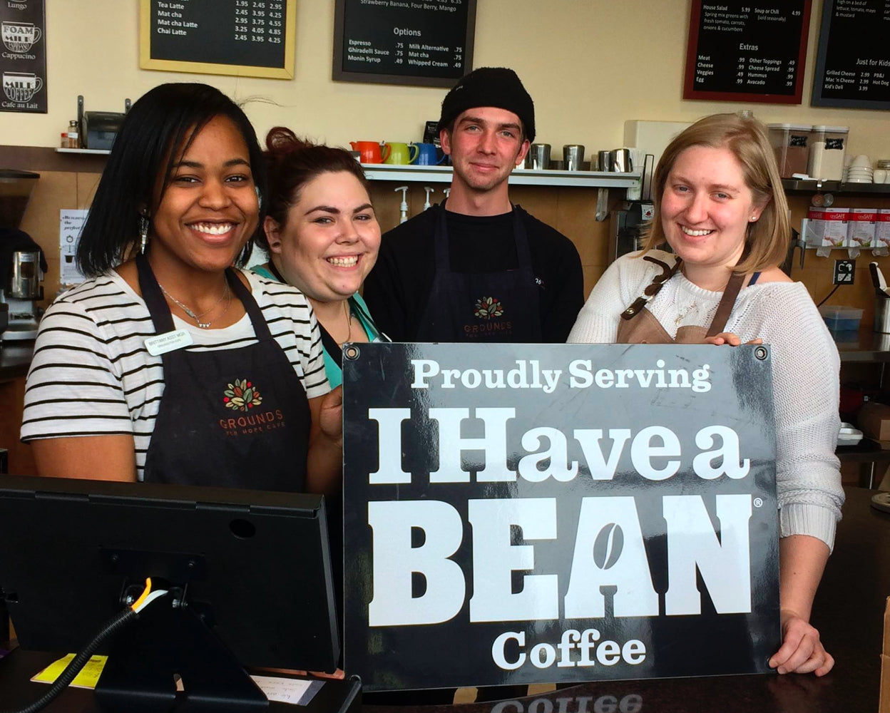 Four baristas from Grounds for Hope Café, three women and one man, holding a sign that reads 'Proudly Serving I Have a Bean Coffee'