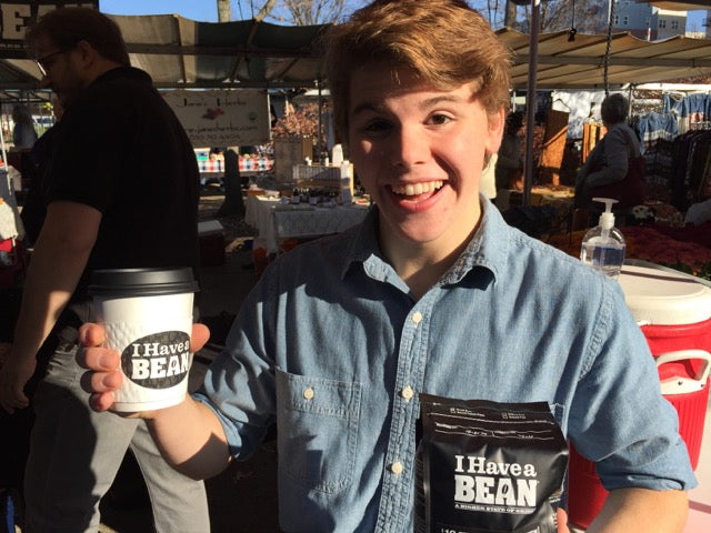 Young white man in a blue button-up shirt holding a white to-go cup and a black coffee bag, both featuring the I Have a Bean logo
